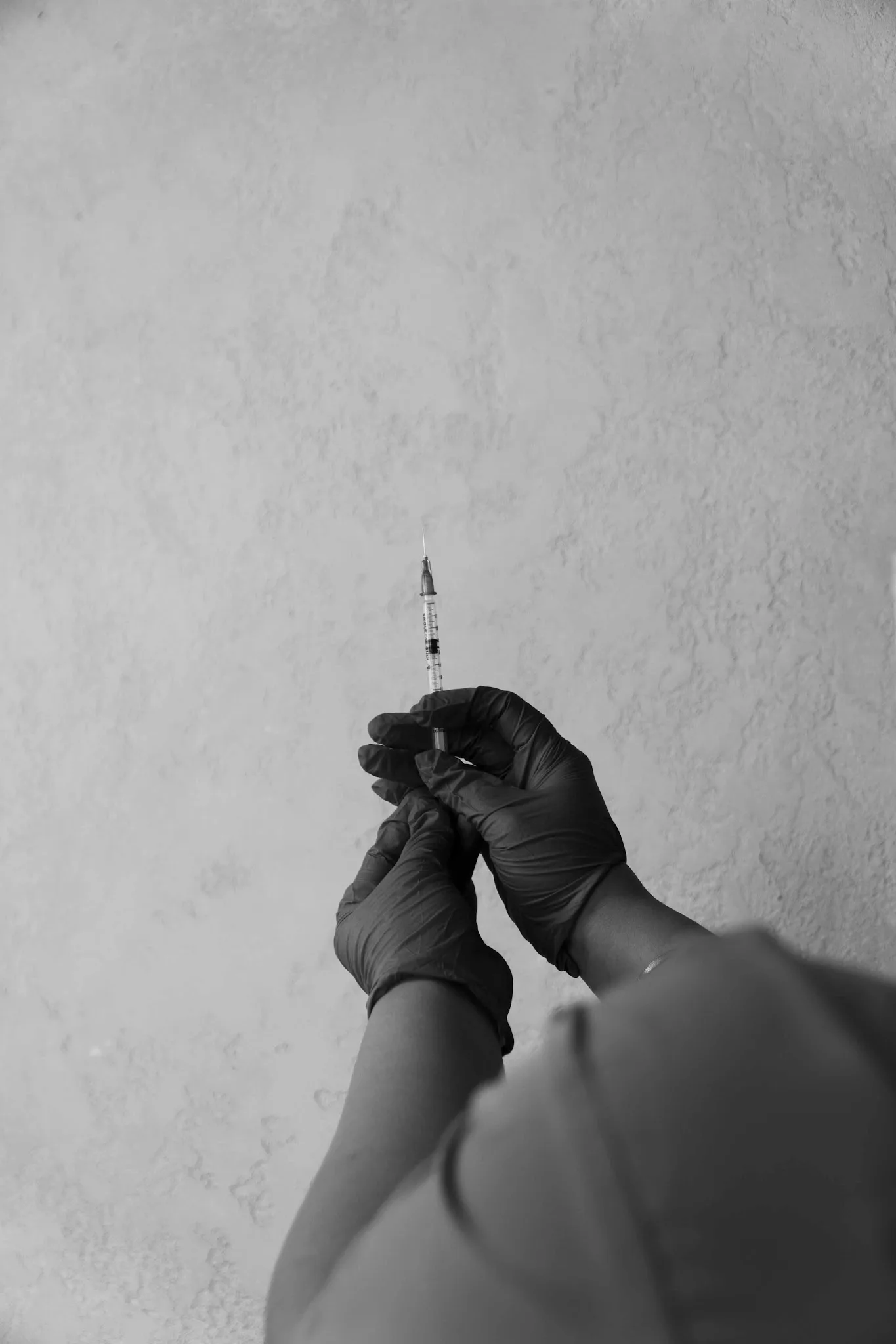 A black-and-white artistic shot of gloved hands holding an aesthetic injection syringe against a textured wall, conveying clinical precision and professionalism.