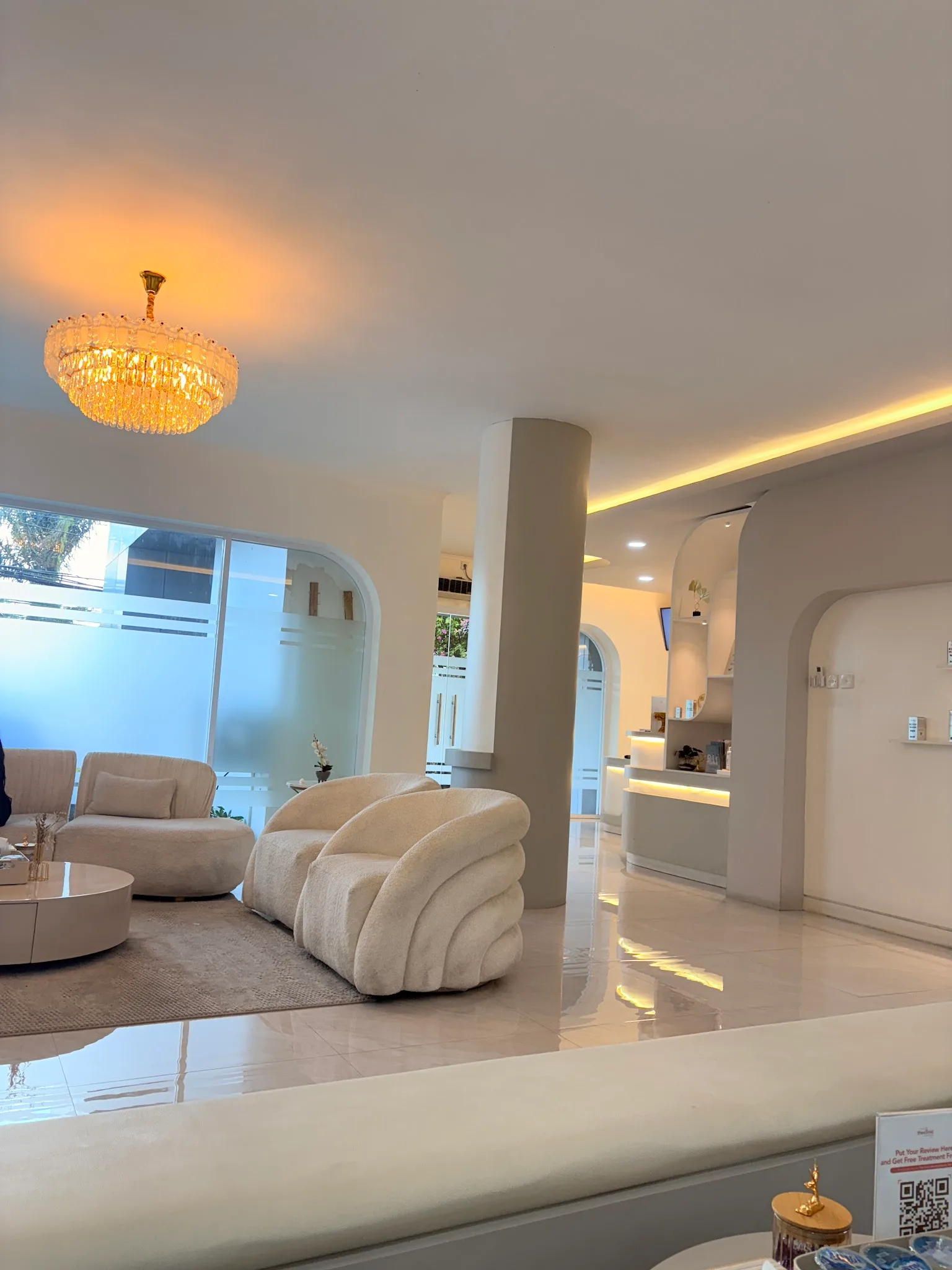 Spacious clinic lobby with ambient warm LED ceiling cove lighting, white columns, built-in wall shelving, seating area, and glossy marble floors. Clean and modern feel.